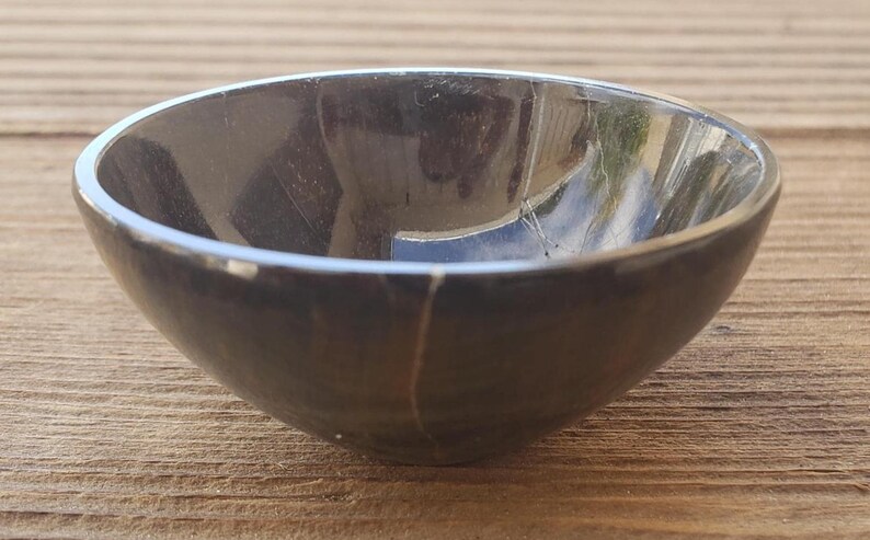 May include: A small, dark gray bowl with a rounded shape. The bowl has a smooth, reflective surface and appears to be made of stone or a similar material. The bowl is sitting on a wooden surface.