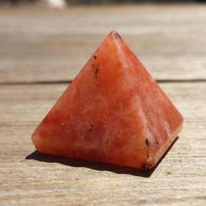 May include: A small, orange-red crystal pyramid with black flecks throughout. The pyramid is sitting on a wooden surface.