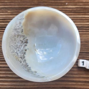 May include: A small, round, white bowl with a light brown and gray pattern on the side. The bowl is sitting on a wooden surface. A white die with the number 15 is next to the bowl.