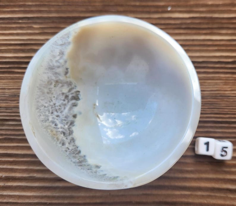 May include: A small, round, white bowl with a light brown and gray pattern on the side. The bowl is sitting on a wooden surface. A white die with the number 15 is next to the bowl.