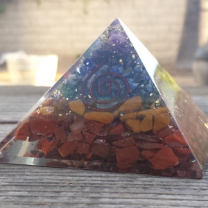 Op de afbeelding: Een heldere glazen piramide gevuld met kleurrijke kristallen en een rood spiraalontwerp. De piramide is gemaakt van helder glas en heeft een verscheidenheid aan kristallen die erin zijn ingebed, waaronder rood, oranje, geel, groen, blauw en paars. De kristallen zijn in een spiraalvormig patroon gerangschikt, met een rood spiraalontwerp in het midden.