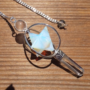 May include: A silver pendulum with a clear crystal point and a star-shaped opal pendant. The pendulum is suspended from a silver chain with a clear crystal bead and a brown crystal bead.