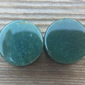 May include: Two round, polished green stones with a glossy finish. The stones are a deep, forest green color and appear to be made of a natural material. They are set against a wooden background.