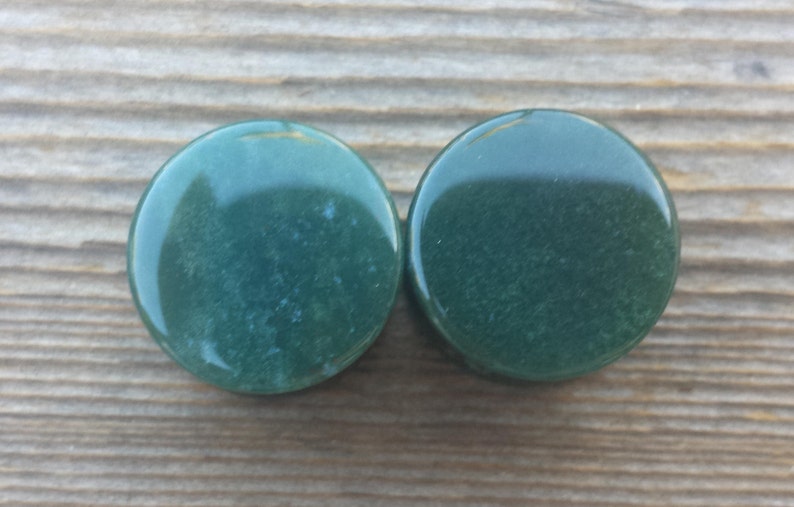 May include: Two round, polished green stones with a glossy finish. The stones are a deep, forest green color and appear to be made of a natural material. They are set against a wooden background.