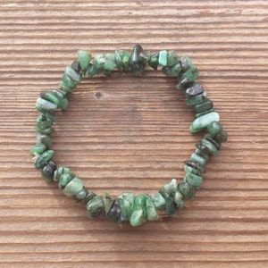 May include: A green gemstone chip bracelet made with small, irregular pieces of emerald. The bracelet is strung on a clear elastic cord and is approximately 7 inches in length.