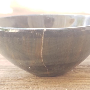 May include: A dark gray, round bowl with a crack running vertically down the side. The bowl has a smooth, polished surface and sits on a wooden surface. The bowl is empty and appears to be made of stone or a similar material.