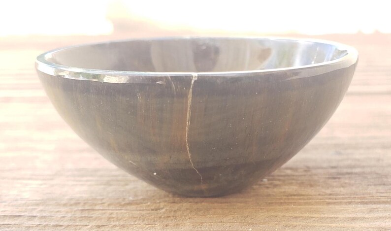 May include: A dark gray, round bowl with a crack running vertically down the side. The bowl has a smooth, polished surface and sits on a wooden surface. The bowl is empty and appears to be made of stone or a similar material.