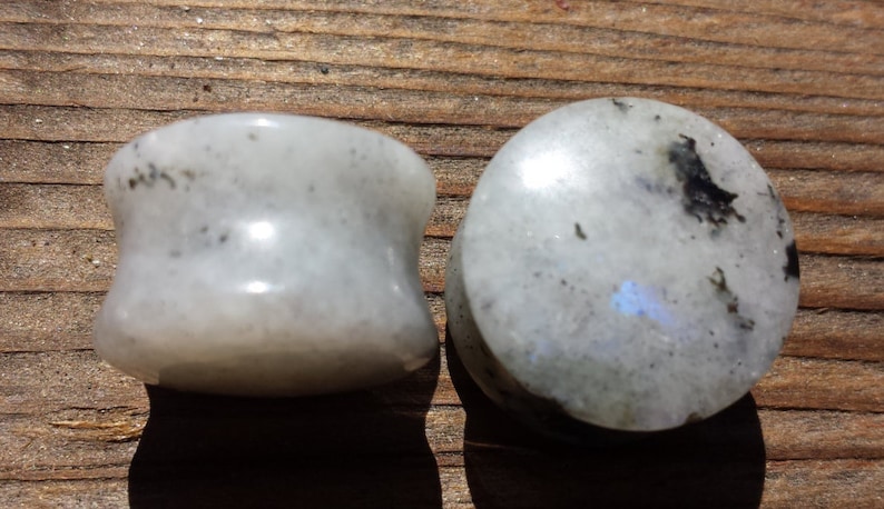 May include: Two white moonstone plugs with black flecks. The plug on the left is a flared style, while the plug on the right is a flat, round style.