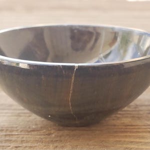 May include: A dark gray bowl with a silver rim. The bowl has a crack running down the side. The bowl is sitting on a wooden surface. The bowl is round and has a smooth surface.