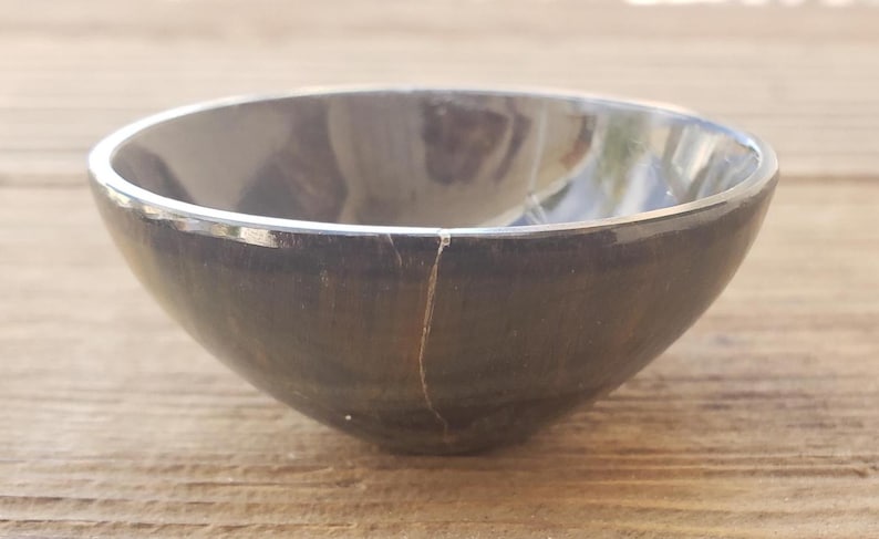 May include: A dark gray bowl with a silver rim. The bowl has a crack running down the side. The bowl is sitting on a wooden surface. The bowl is round and has a smooth surface.