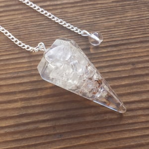 May include: A clear quartz crystal pendulum with a silver chain. The crystal is faceted and has a point at the bottom. The chain is attached to a small silver ring at the top of the crystal.