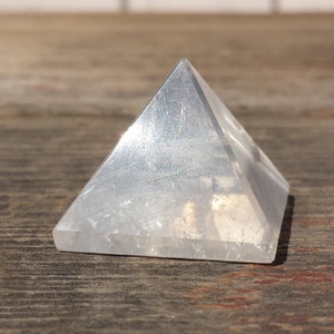 May include: A clear crystal pyramid, with a smooth, polished surface. The pyramid is sitting on a wooden surface.