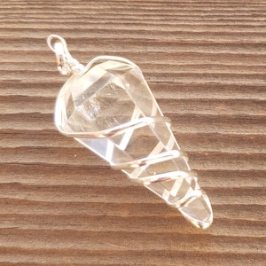 May include: A clear quartz crystal point wrapped in silver wire, creating a pendant. The pendant is on a brown wooden surface.