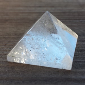 May include: A clear quartz crystal pyramid on a wood surface. The pyramid is made of clear quartz and has a smooth, polished finish.