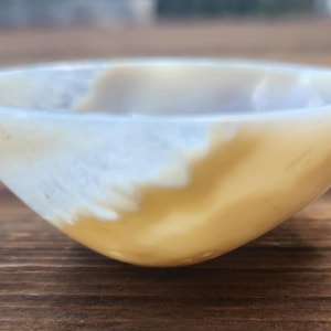 May include: A small, handcrafted bowl with a smooth, polished surface. The bowl features a gradient of colors, transitioning from a creamy white to a warm, golden-yellow hue. The bowl's shape is round and shallow, perfect for holding small items.