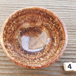 Pu&ograve; includere: Una piccola ciotola rotonda marrone con interno lucido ed esterno strutturato. La ciotola &egrave; realizzata con un materiale naturale, forse pietra o minerale. La ciotola &egrave; appoggiata su una superficie di legno. I numeri 4 e 7 sono su un cubo bianco.