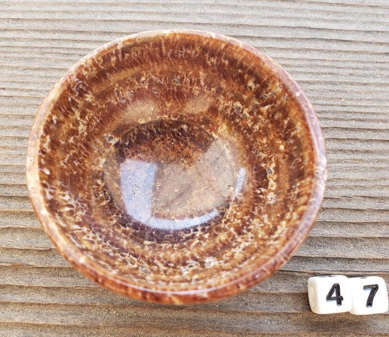 Pu&ograve; includere: Una piccola ciotola rotonda marrone con interno lucido ed esterno strutturato. La ciotola &egrave; realizzata con un materiale naturale, forse pietra o minerale. La ciotola &egrave; appoggiata su una superficie di legno. I numeri 4 e 7 sono su un cubo bianco.