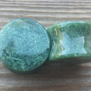 May include: Two green stone plugs with a polished finish. The plugs have a rounded shape with a slight concave curve on the sides. The stone has a mottled pattern of lighter and darker green.
