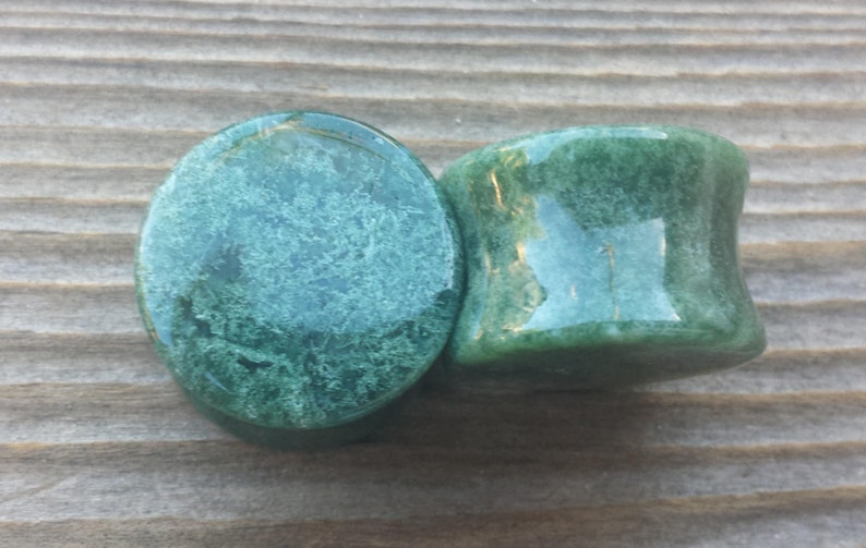 May include: Two green stone plugs with a polished finish. The plugs have a rounded shape with a slight concave curve on the sides. The stone has a mottled pattern of lighter and darker green.