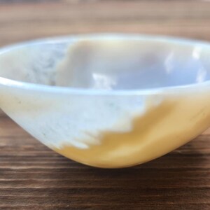 May include: A small, round bowl with a smooth, polished surface. The bowl features a blend of creamy white and golden-yellow hues, creating a marbled effect. The bowl's interior is a lighter shade, and it sits on a wooden surface.