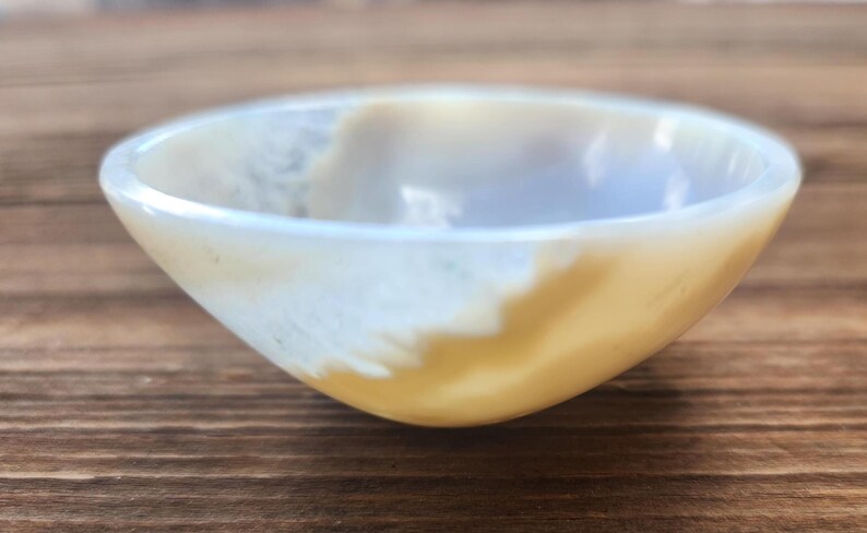May include: A small, round bowl with a smooth, polished surface. The bowl features a blend of creamy white and golden-yellow hues, creating a marbled effect. The bowl's interior is a lighter shade, and it sits on a wooden surface.