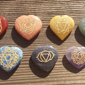 May include: Seven heart-shaped stones with gold chakra symbols engraved on them. The stones are arranged in a row from left to right, with the colors red, orange, yellow, green, blue, indigo, and violet.