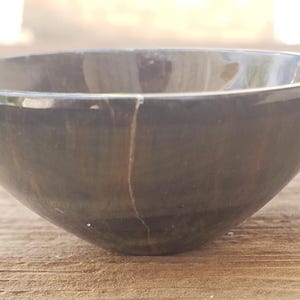 May include: A dark gray, smooth bowl with a rounded shape. The bowl has a crack running down the side. The bowl is sitting on a wooden surface.