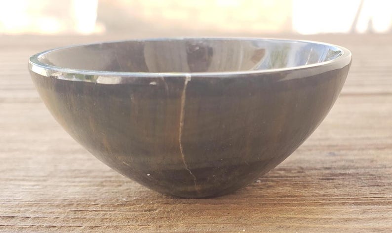 May include: A dark gray, smooth bowl with a rounded shape. The bowl has a crack running down the side. The bowl is sitting on a wooden surface.