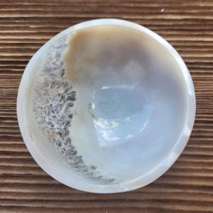 May include: A polished, round bowl with a natural, organic design. The bowl features a blend of creamy white, tan, and gray tones, with a textured, wave-like pattern along the edge. The interior has a smooth, glossy finish.