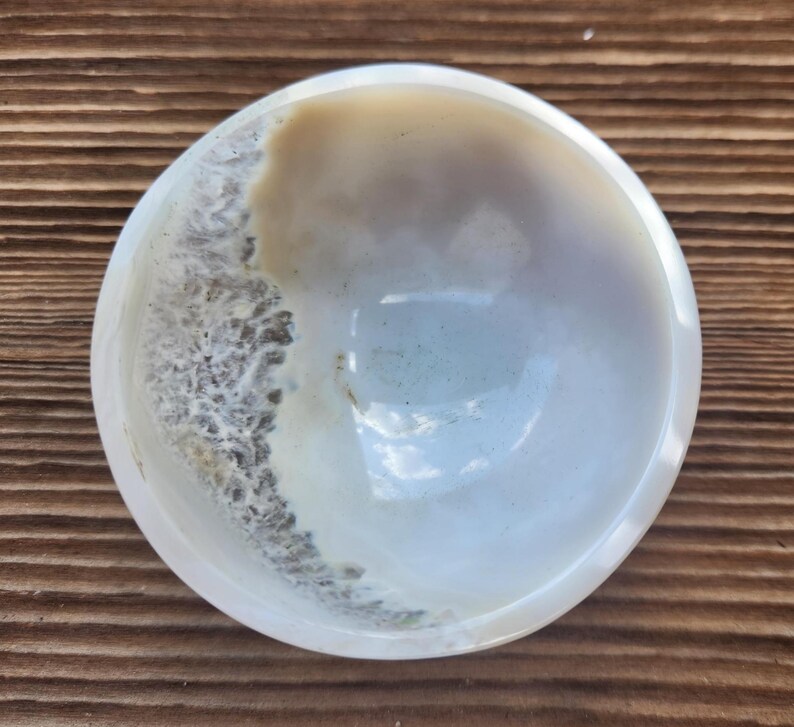 May include: A polished, round bowl with a natural, organic design. The bowl features a blend of creamy white, tan, and gray tones, with a textured, wave-like pattern along the edge. The interior has a smooth, glossy finish.