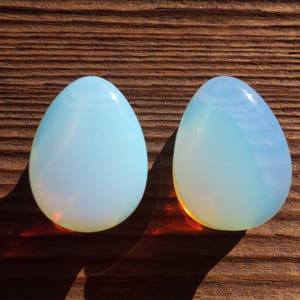 May include: Two teardrop-shaped, iridescent opal stones on a wooden surface. The stones are a pale blue color with a slight shimmer.