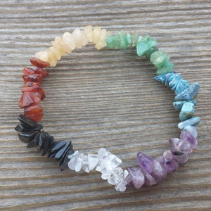 May include: A rainbow chakra bracelet made with small, chipped gemstones in various colors, including red, orange, yellow, green, blue, indigo, and violet. The bracelet is laid out on a wooden surface.