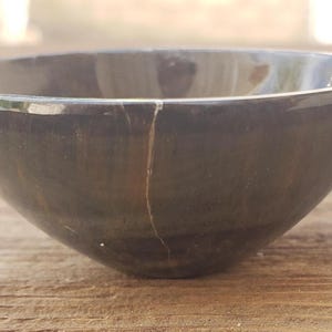 May include: A dark gray, round bowl with a smooth, polished surface. The bowl has a crack running down the side. The bowl is made of a dark stone material and is sitting on a wooden surface.
