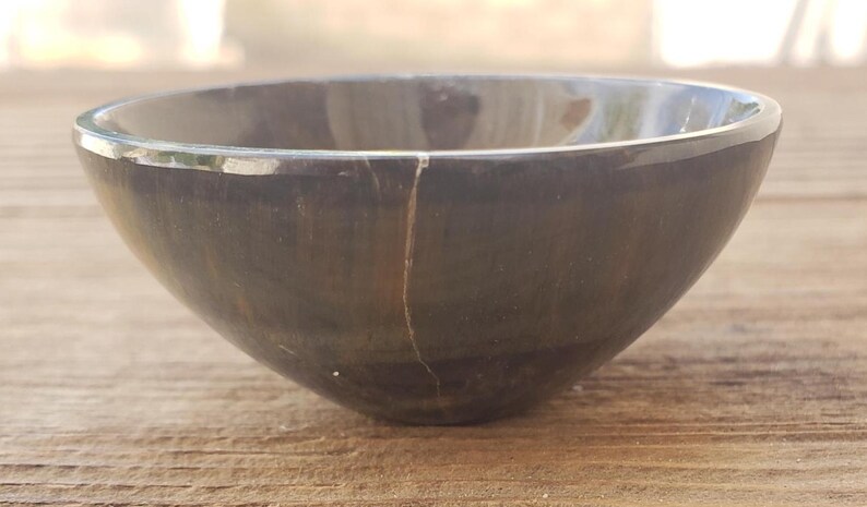 May include: A dark gray, round bowl with a smooth, polished surface. The bowl has a crack running down the side. The bowl is made of a dark stone material and is sitting on a wooden surface.