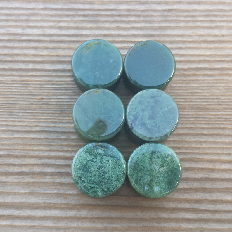 May include: Six round, polished green stone plugs are arranged on a wooden surface. The plugs vary in texture, with some appearing smooth and others showing a mossy, speckled pattern. The color ranges from deep forest green to lighter shades.