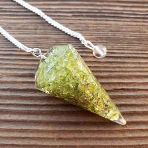 May include: A green crystal pendulum with a silver chain and a clear crystal bead at the end. The pendulum is on a wooden surface.