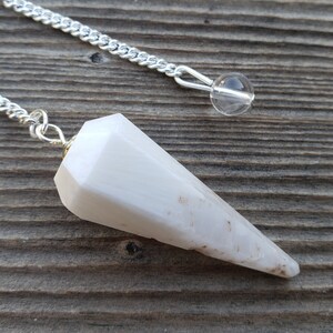May include: White crystal pendulum with a silver chain and a clear crystal bead. The pendulum is pointed and has a faceted design.