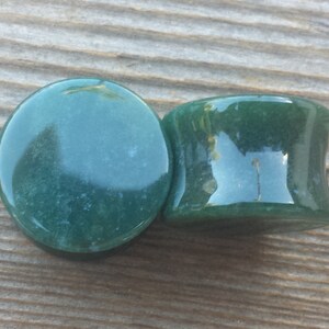May include: Two dark green stone ear plugs. One plug is flat and round, while the other has a flared, cylindrical shape. The plugs are smooth and polished, with slight variations in color and texture. The plugs are made of a natural stone material.