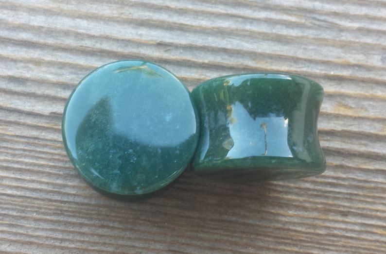 May include: Two dark green stone ear plugs. One plug is flat and round, while the other has a flared, cylindrical shape. The plugs are smooth and polished, with slight variations in color and texture. The plugs are made of a natural stone material.