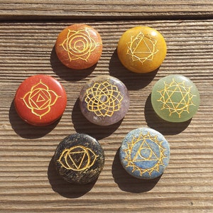 May include: Seven chakra stones with gold symbols on a wooden surface. The stones are red, orange, yellow, green, blue, indigo, and violet.