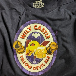 Puede incluir: Una camiseta morada y amarilla con un gráfico de una etiqueta de cerveza. La etiqueta dice "Wily Castle Yellow Devil Ale" y presenta un personaje de dibujos animados sosteniendo una jarra de cerveza. La etiqueta también incluye un pequeño gráfico de castillo y el texto "1987 Awarded to Albright in Wily Castle Brewmaster."