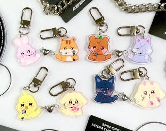 Pode incluir: Um conjunto de oito chaveiros coloridos com personagens de animais de desenho animado. Cada chaveiro tem um anel de metal prateado e um amuleto de acrílico transparente com um design de animal diferente. Os animais incluem um coelho rosa, um esquilo castanho, um urso castanho, um gato roxo, um pintinho amarelo, um cão amarelo, um gato azul e um cão branco. Os chaveiros estão todos dispostos numa superfície branca com um fundo preto.