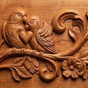 May include: A detailed wood carving of two birds nestled together, with a heart shape between them. The birds are perched on a branch with leaves and flowers. The carving is made of a warm, light brown wood.