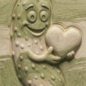 May include: A carved wooden relief depicting a smiling pickle character holding a heart. The pickle is green with a textured surface and the heart is a lighter color. The background has a striped pattern.