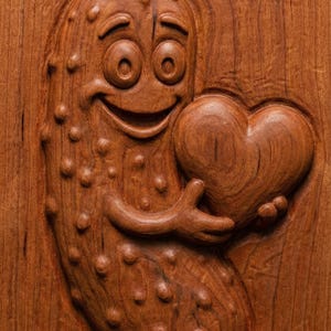 May include: A wooden carving of a smiling pickle character holding a heart. The pickle has a happy expression and is covered in small bumps. The heart is smooth and detailed, and the carving is set against a wood grain background.