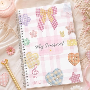 May include: Spiral-bound journal with a pink and white gingham pattern. The cover features fabric embellishments including a bow, flowers, stars, and hearts. The words "My Journal" are written on the cover. A gold pen and decorative elements surround the journal.