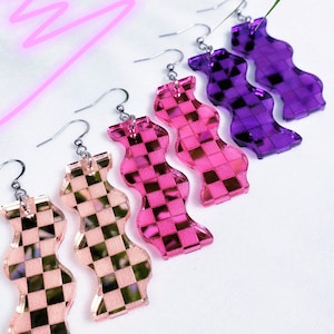 May include: A selection of colourful, wavy-shaped earrings in shades of pink, purple, and peach. Each earring has a checkered pattern and a silver hook. The earrings are displayed on a white surface with a neon pink squiggle.
