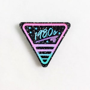 May include: A black triangle-shaped pin with pink and blue glitter and a white outline. The pin has the text "1980s" in blue and purple lettering.