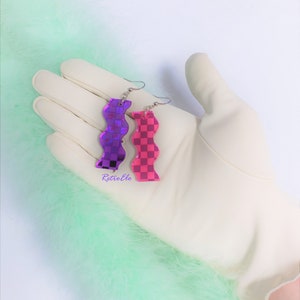 May include: A pair of wavy earrings, one purple and one pink, with a checkered pattern. The earrings are shown on a white gloved hand, set against a backdrop of light green feathers. Silver hooks.