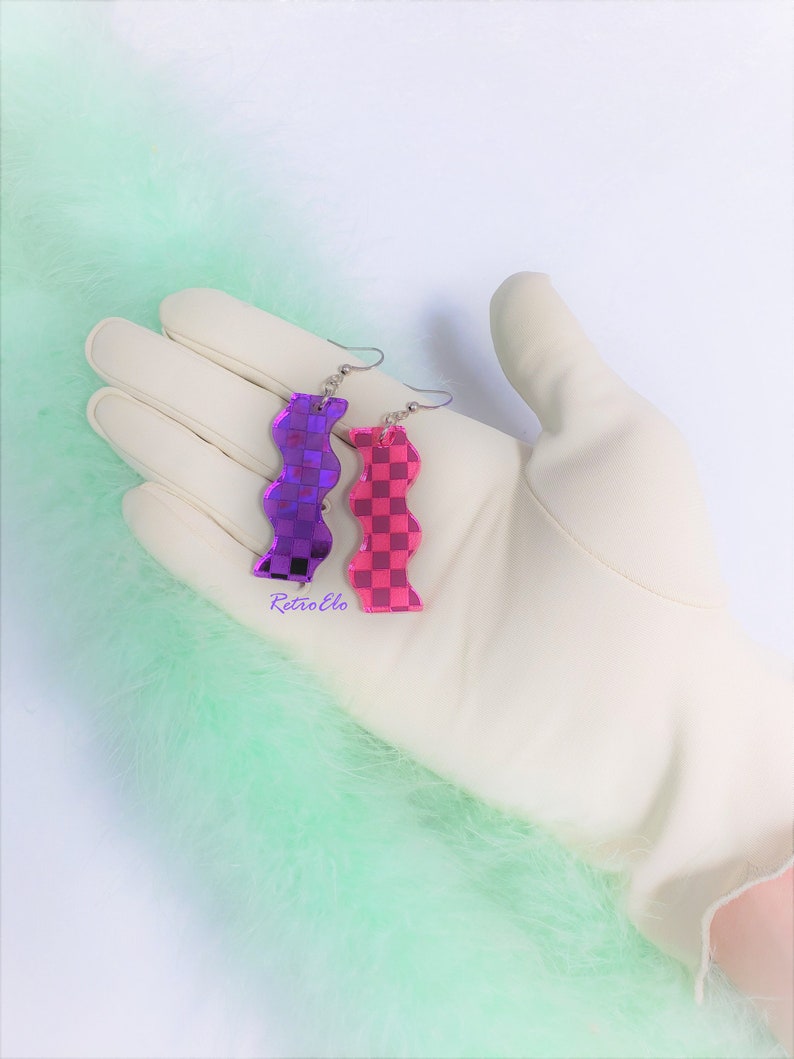 May include: A pair of wavy earrings, one purple and one pink, with a checkered pattern. The earrings are shown on a white gloved hand, set against a backdrop of light green feathers. Silver hooks.